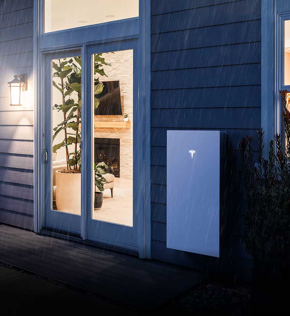Tesla-Battery-Storage-03 Wall-mounted home battery storage unit on the exterior of a house beside glass doors